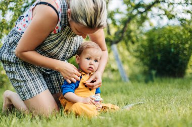 Genç bir anne, küçük oğluyla bahçede çimlerin üzerinde oturur ve elbiselerini düzeltir..