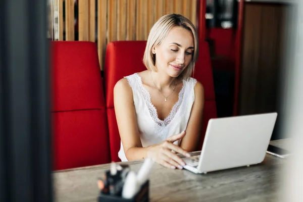 Uma linda garota loira usa um laptop e trabalha em um café. Pausa, trabalho online, freelance — Fotografia de Stock