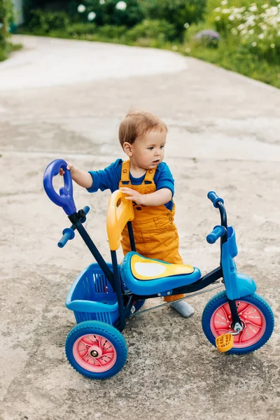 Um rapazinho está ao lado de um triciclo infantil. Primeira bicicleta, novas habilidades — Fotografia de Stock