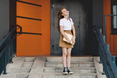 Sırt çantalı bir kız öğrenci evin yanındaki merdivenlerde duruyor, elinde bir defter, temiz havanın tadını çıkarıyor, üniversiteye gidiyor.