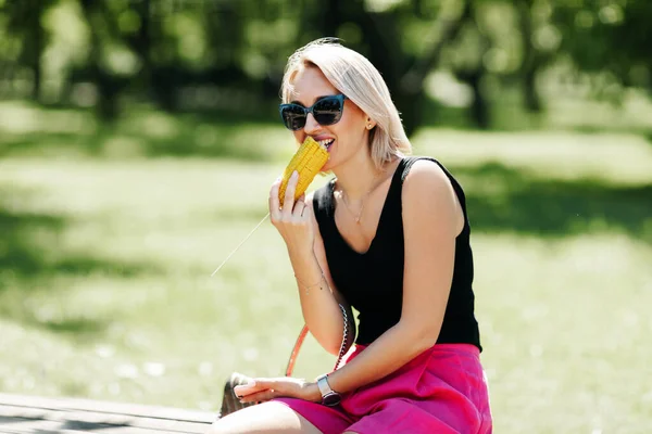 Blondýnka sedí na lavičce v parku a jí horkou kukuřici, odpočívá po procházce — Stock fotografie