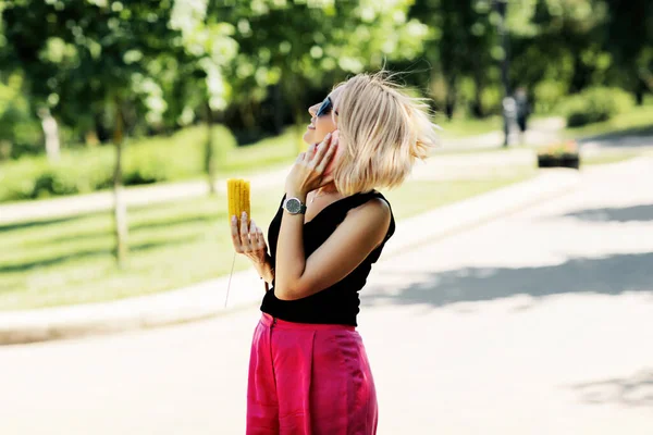 Mladá žena ve slunečních brýlích jí kukuřici a telefonuje v letním parku, což je pojetí jídla pro cestování — Stock fotografie