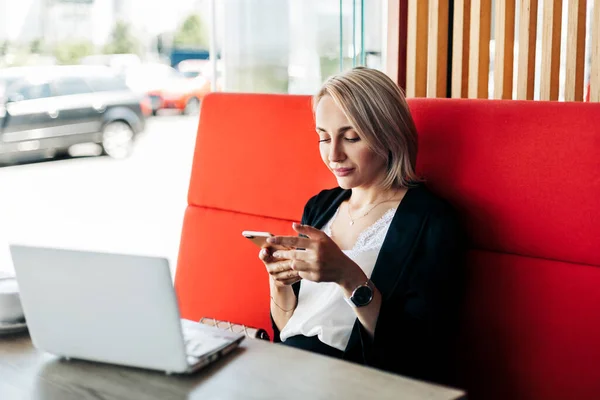 Uma mulher autônoma está trabalhando com seu telefone e laptop no terraço de um restaurante sobre uma xícara — Fotografia de Stock