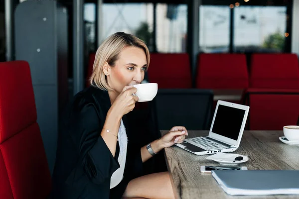 Uma jovem está bebendo café em um café durante uma pausa e trabalhando em um laptop. Conceito de negócio — Fotografia de Stock