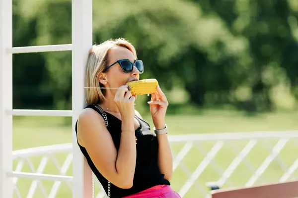 Krásná dívka ve slunečních brýlích stojí u altánku v parku a jí horkou kukuřici. Léto, dovolená, park — Stock fotografie