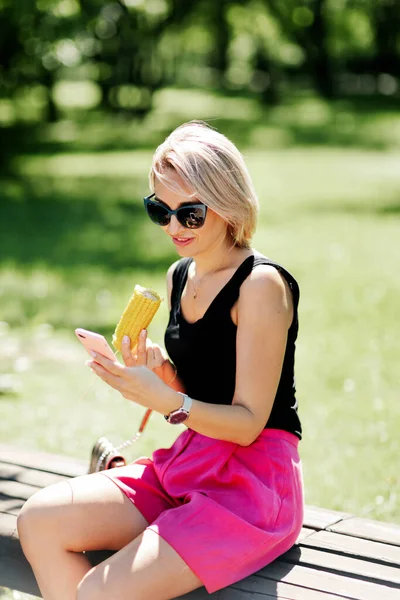 Blondýnka sedí na lavičce v parku a jí horkou kukuřici, odpočívá po procházce — Stock fotografie