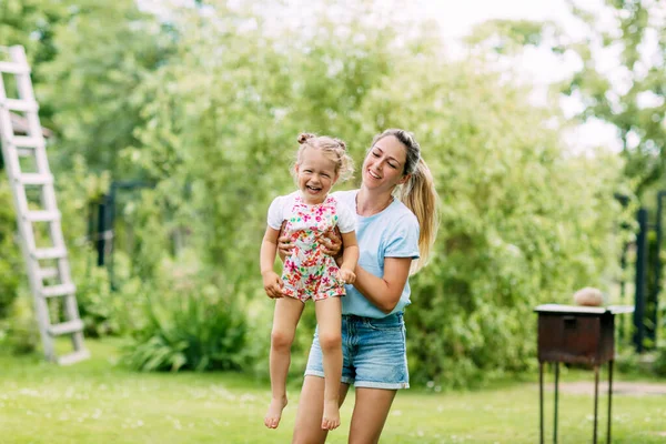 Uma jovem mãe e sua linda garotinha estão se divertindo em um jardim ensolarado. O conceito de uma infância feliz e maternidade, uma mãe segura sua filhinha em seus braços — Fotografia de Stock