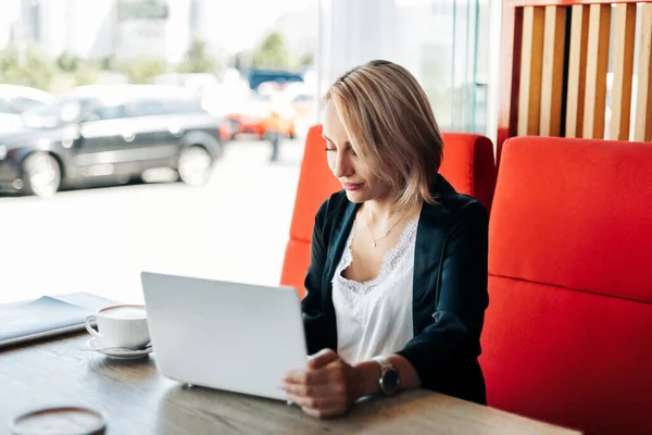 Uma linda garota loira usa um laptop e trabalha em um café. Pausa, trabalho online, freelance — Fotografia de Stock