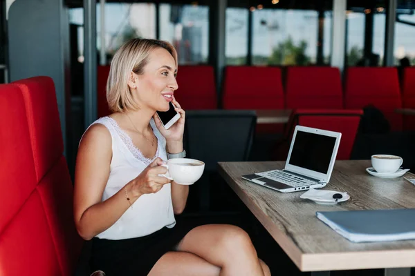 Uma jovem está falando ao telefone e bebendo café em um café, fazendo uma pausa enquanto trabalhava — Fotografia de Stock