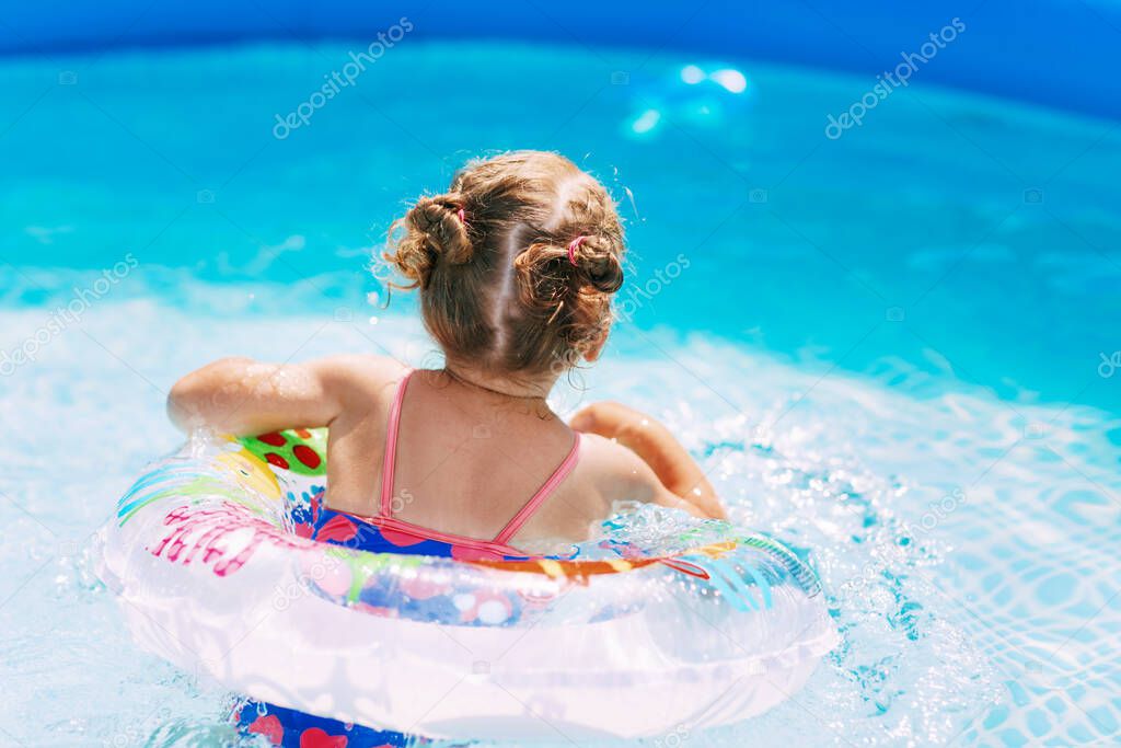 A charming little girl in a bright swimsuit learns to swim in the pool in the garden with the help of an inflatable circle. Summer time, recreation, rear view