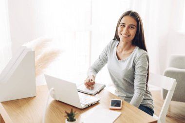 Masada oturan genç bir öğrenci çalışırken dizüstü bilgisayar kullanıyor, not defterine not alıyor.