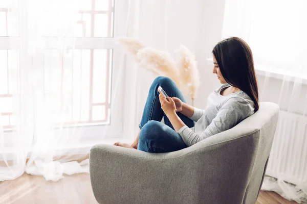 Uma linda garota se senta em uma cadeira com um telefone nas mãos, escreve mensagens, faz compras, visualizações e publica posts nas redes sociais. Mídia social, comunicação online — Fotografia de Stock