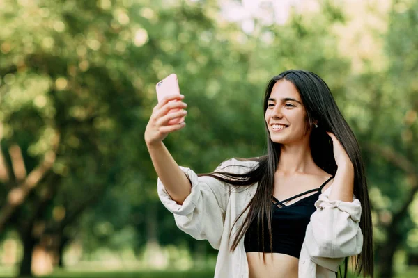 Una joven con el pelo castaño largo toma una selfie en un teléfono inteligente en el parque — Foto de Stock