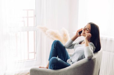 Genç bir kız cep telefonuyla konuşuyor ve sandalyeye oturmuş ayaklarıyla ona tırmanıyor.