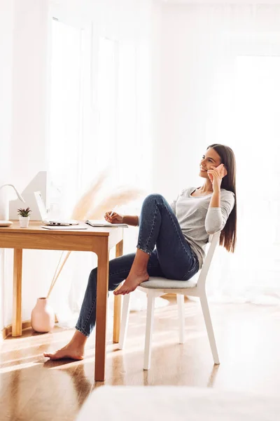 Uma jovem está falando ao telefone em sua mesa, usando um laptop., fazendo anotações em um caderno — Fotografia de Stock