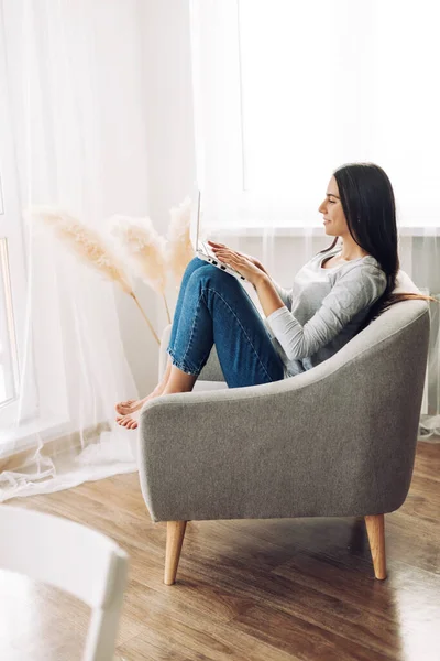 Uma menina bonita com um laptop está sentada em uma cadeira macia e confortável e trabalhando em um laptop, digitando texto. Tecnologias modernas, redes sociais — Fotografia de Stock