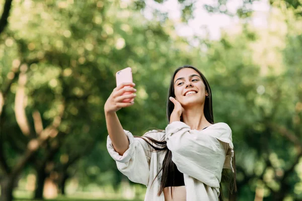 Una joven con el pelo castaño largo toma una selfie en un teléfono inteligente en el parque — Foto de Stock