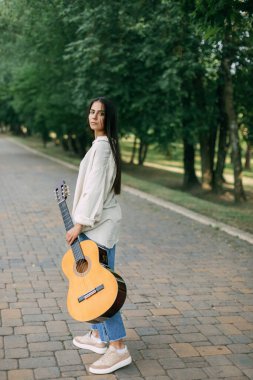 Akustik gitarlı güzel siyah saçlı bir kız parkta bir ara sokakta yürüyor.