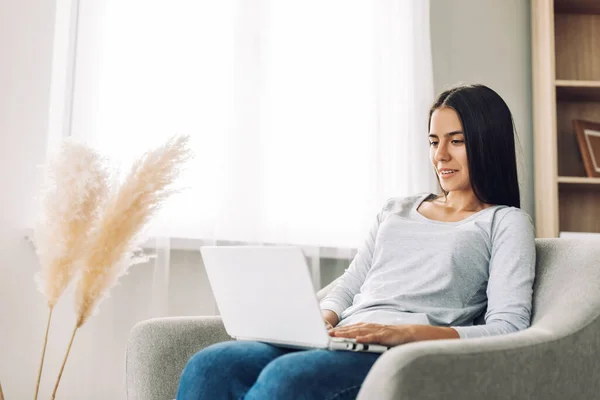 Seorang gadis muda belajar menggunakan laptop, menonton video kuliah, duduk di kursi di rumah, berkomunikasi dengan teman-teman — Stok Foto