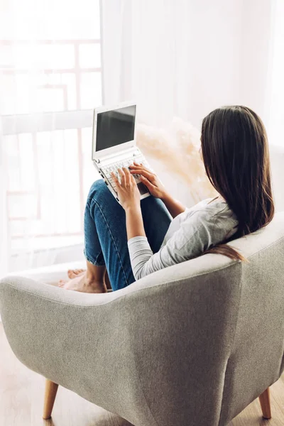 노트북을 들고 있는 예쁜 소녀가 부드러운 편안 한 의자에 앉아서 노트북 컴퓨터로 글을 쓰며 일하고 있습니다. 현대의 기술, 소셜 미디어 — 스톡 사진