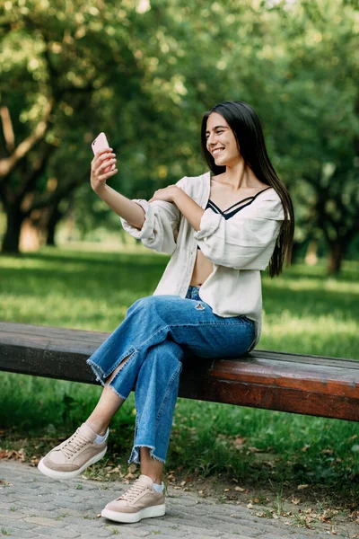 Una atractiva morena alegre con el pelo largo se toma emocionalmente una selfie sentada en un banco del parque. El concepto de unas vacaciones relajantes — Foto de Stock