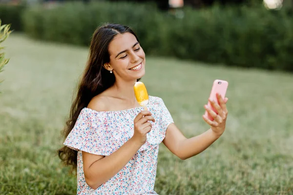 Una chica está sentada en la hierba del parque, comiendo helado y sosteniendo un teléfono en sus manos. — Foto de Stock