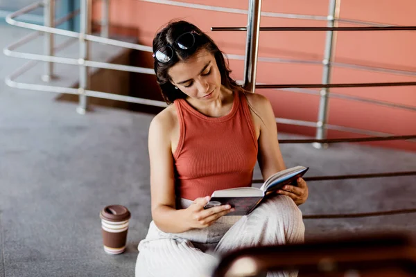 Una estudiante se sienta en las escaleras del campus y lee un libro, junto a ella hay un vaso de café —  Fotos de Stock