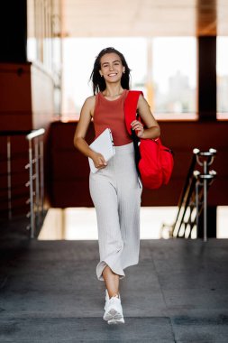 Sırt çantası ve dizüstü bilgisayarı olan gülümseyen bir öğrenci üniversitede merdivenlerden çıkıp derslere koşuyor.