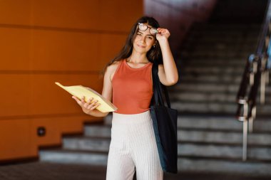 Elinde dosya olan güzel bir esmer üniversitede ya da üniversitede merdivenlerin yanında duruyor.