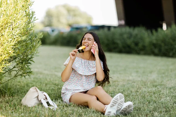 Uma linda garota está falando ao telefone, sentada na grama e comendo sorvete. — Fotografia de Stock