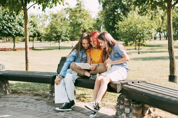 Tři půvabné školačky sedí na lavičce v parku a připravují se na společné lekce nebo zkoušky. Přátelé, přátelství, společný čas — Stock fotografie