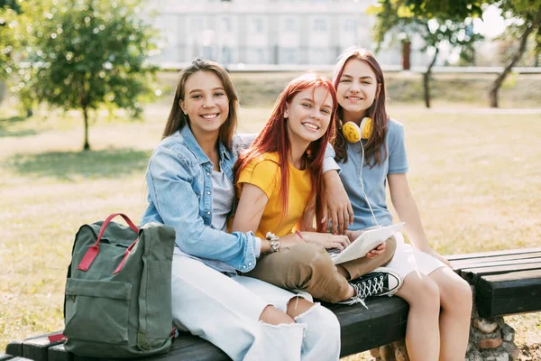 Três alunas estão sentadas em um banco do parque, conversando, preparando-se junto com a lição. O conceito de formação e educação — Fotografia de Stock