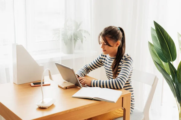 A dark-haired schoolgirl sits at a desk doing her homework using her tablet. Modern technologies, online education, training — Fotografia de Stock