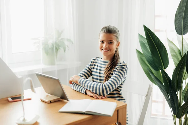 Smiling schoolgirl in a striped sweater sits at the table, does homework on the tablet and takes notes in a notebook. Modern technologies, online education, training — Stock Photo, Image