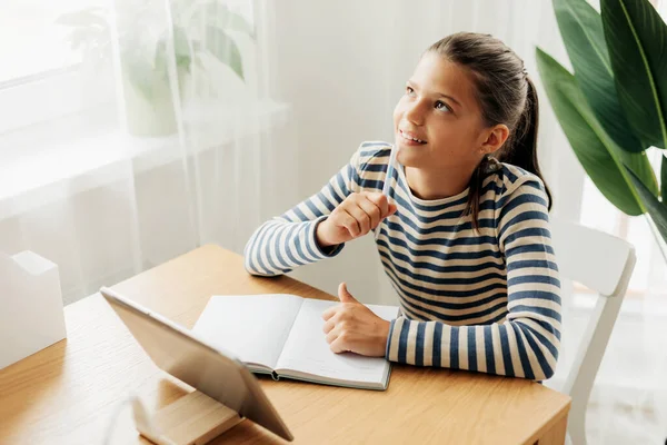 Happy pensive schoolgirl sits at the table at home, does homework on the tablet and looks out the window. Modern technologies, online education, training — Fotografia de Stock