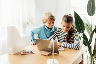 Küçük erkek kardeşiyle aynı masada oturan bir kız öğrenci bir tablet üzerinde ödev yapar ve bir deftere not alır.