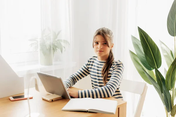 A serious schoolgirl in a striped sweater is sitting at a table, doing her homework on her tablet and taking notes in a notebook. Modern technologies, online education, training — Stock Photo, Image