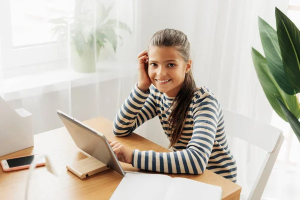 Smiling schoolgirl in a striped sweater sits at the table, does homework on the tablet and takes notes in a notebook. Modern technologies, online education, training — Fotografia de Stock