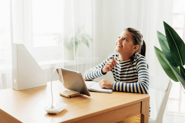 Happy pensive schoolgirl sits at the table at home, does homework on the tablet and looks out the window. Modern technologies, online education, training — Stock Photo, Image
