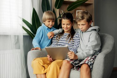 Güzel siyah saçlı kız kardeş ve erkek kardeşleri evde kanepede oturuyorlar, tablet üzerinde oyunlar oynuyorlar ve video aracılığıyla arkadaşlarıyla sohbet ediyorlar. Modern teknoloji ve evde zaman geçirme