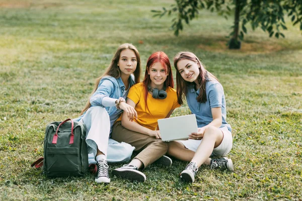 Las adolescentes están sentadas en un césped verde en un parque con una mochila y una tableta digital y están preparando un proyecto conjunto. Educación y conocimiento, estilo de vida de los adolescentes — Foto de Stock