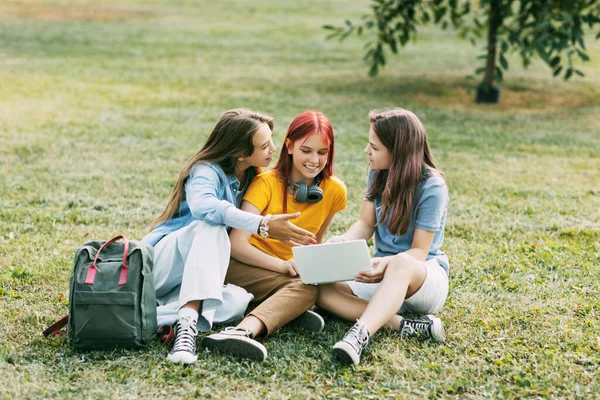 Las adolescentes están sentadas en un césped verde en un parque con una mochila y una tableta digital y están preparando un proyecto conjunto. Educación y conocimiento, estilo de vida de los adolescentes — Foto de Stock