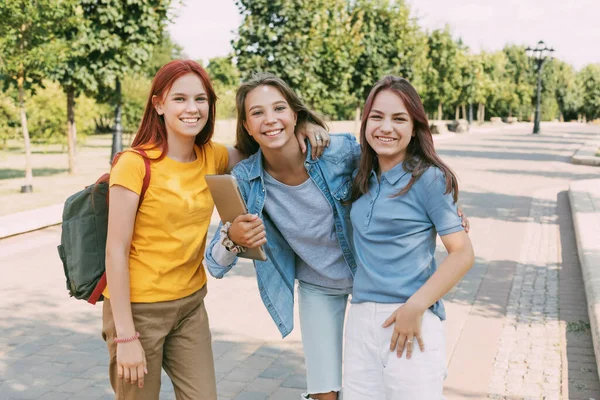 Tre tonårstjejer går med en surfplatta och en ryggsäck längs parkens gränd till skolan. Begreppet utbildning. Tillbaka till skolan — Stockfoto