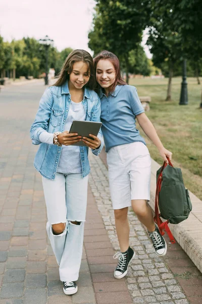 Dos colegialas están sosteniendo una tableta en sus manos y viendo un video, riéndose en el camino a la escuela. Amigos, amistad, tiempo juntos — Foto de Stock