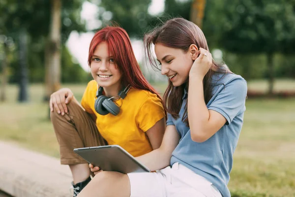 Dos amigas colegialas encantadoras están sentadas en el parque después de la escuela y viendo videos en una tableta o preparando un proyecto conjunto. — Foto de Stock