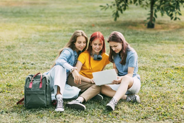 Las adolescentes están sentadas en un césped verde en un parque con una mochila y una tableta digital y están preparando un proyecto conjunto. Educación y conocimiento, estilo de vida de los adolescentes — Foto de Stock