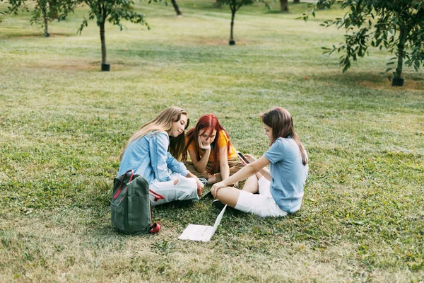 Teenage dívky sedí na zeleném trávníku v parku s batohem a digitálním tabletem a připravují společný projekt. Vzdělání a znalosti, životní styl teenagerů — Stock fotografie
