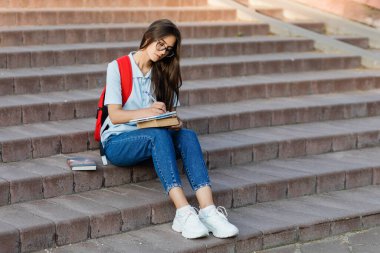Gözlüklü, merdivenlerde oturan odaklanmış bir öğrenci defterine bir şeyler yazıyor. Ders ya da sınav hazırlığı