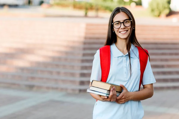 Una estudiante sonriente con gafas, libros y una mochila roja. Retrato de un estudiante en un fondo de la ciudad. El concepto de educación — Foto de Stock