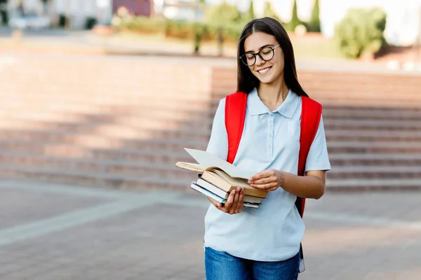 Una chica encantadora con una mochila y gafas está leyendo un libro. Retrato de un estudiante en un fondo de la ciudad. El concepto de educación — Foto de Stock